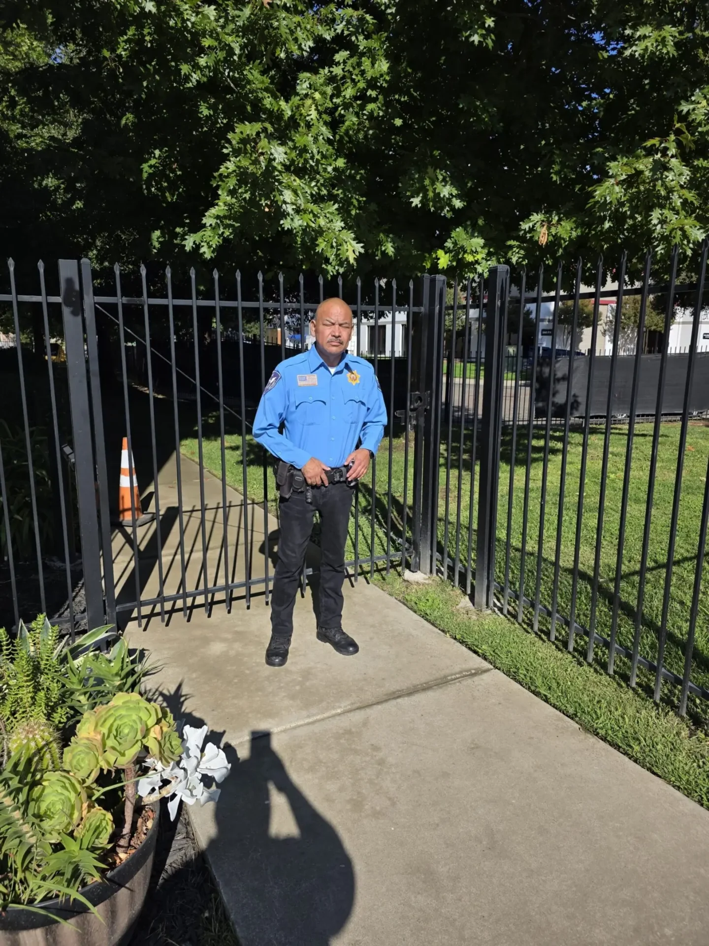 Security guard standing at an open gate near a grassy area with trees.